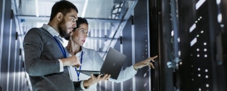 Zwei IT Techniker stehen vor einem Server und besprechen die Ergebnisse der Wartung auf deren Laptop.