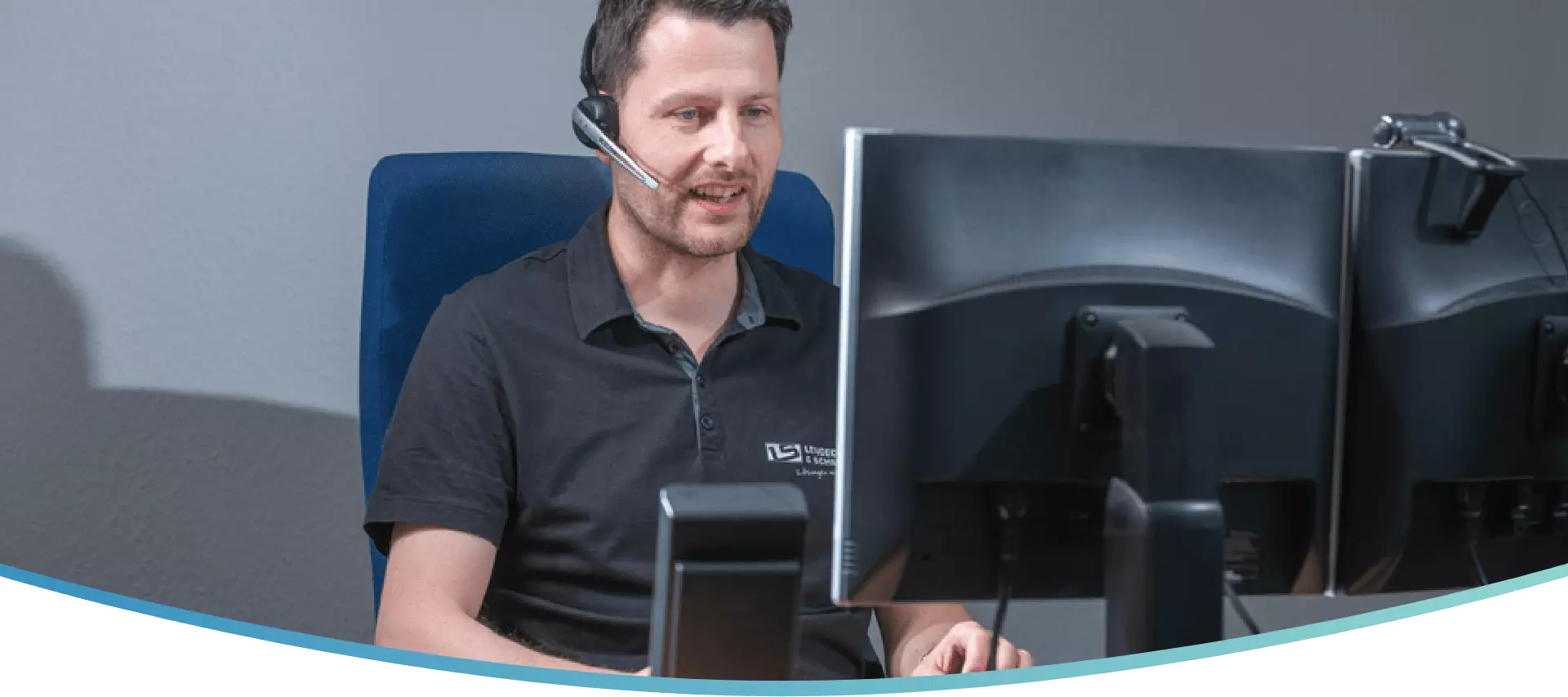 Ein IT Techniker sitzt an seinem Computer und telefoniert mit seinem Headset mit einem Kunden.
